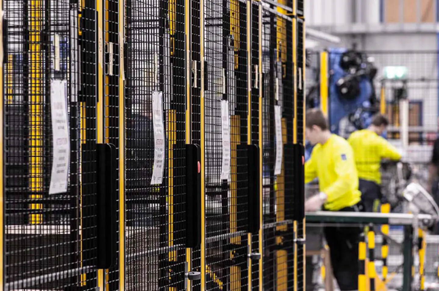 Workers in a factory, next to black and yellow safety barriers with metal mesh panels